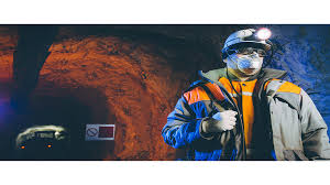 A miner in an underground tunnel wearing a hard hat with a headlamp, mask, and high-visibility safety gear.